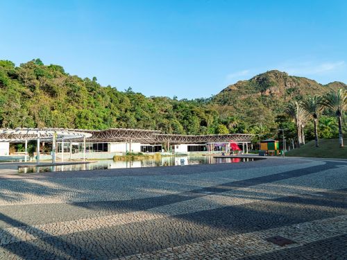 Praça ampla de piso de pedras, com lago e estrutura metálica de cobertura em frente. Ao fundo, encosta arborizada e montanha com vegetação e rochas. Algumas palmeiras à direita.