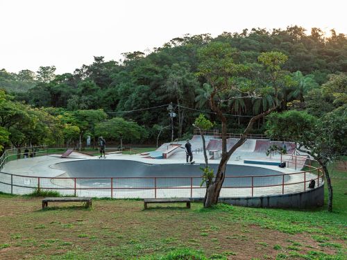 Pista de skate circular em concreto com corrimão metálico vermelho. Dois praticantes estão de pé com skates. Vegetação verde densa ao redor, com árvores grandes.