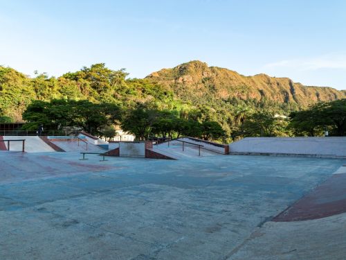 Área de skate com rampas e obstáculos de concreto. Ao fundo, árvores verdes e uma encosta de montanha com vegetação e áreas de rocha exposta. Céu claro.
