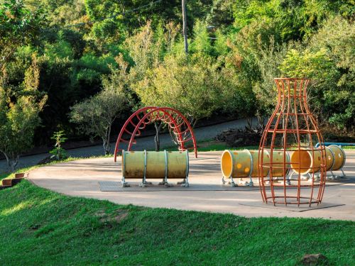 Playground ao ar livre com túneis cilíndricos amarelos sobre suportes metálicos, arco de escalada vermelho e estrutura vazada laranja em forma de garrafa. Piso de concreto e gramado em primeiro plano. Ao fundo, árvores densas, caminho de cascalho, postes e cabos aéreos.