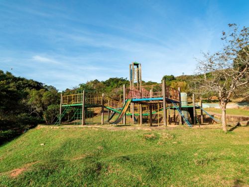 Brinquedo infantil elevado de metal e madeira com passarelas, torres e vários escorregadores verdes e azuis; torre mais alta ao centro. Estrutura sobre morro gramado, árvores ao redor e céu azul. À direita, árvore de galhos finos sem folhas.