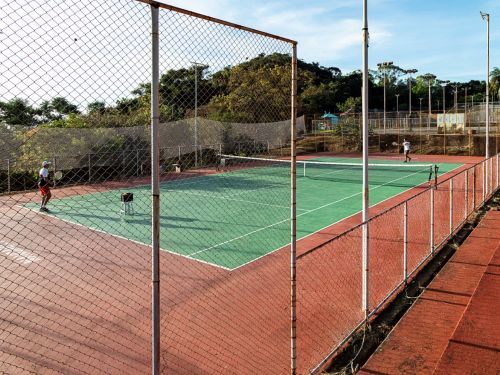 Quadra de tênis cercada por alambrado metálico. O piso é verde no centro e vermelho nas laterais. Dois jogadores estão em lados opostos da rede, com raquetes. Há refletores altos ao redor e vegetação com árvores e morros ao fundo.