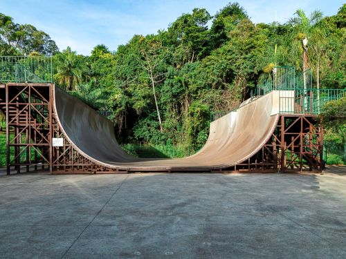 Uma rampa em formato de “U” de skate com plataformas nas extremidades e grades verdes no topo. Estrutura apoiada em treliças sobre piso de concreto. Ao fundo, vegetação densa com árvores altas e palmeiras; céu claro.