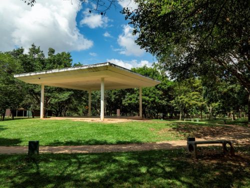 Grande pavilhão aberto no Parque Ecológico da Pampulha. O pavilhão tem um telhado quadrado e plano, sustentado por quatro colunas finas. Ele está em uma área de grama verde, com um caminho de cimento claro ao lado. O parque está cercado por árvores com folhagem densa. O céu é azul com grandes nuvens brancas e esparsas.