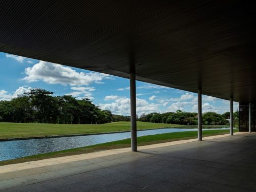 Uma passarela coberta no Parque Ecológico da Pampulha. O teto escuro, sustentado por colunas finas, faz uma moldura para a paisagem. A vista é de um canal de água azul com um gramado verde do outro lado. O horizonte é delimitado por árvores, sob um céu azul com nuvens grandes e brancas.