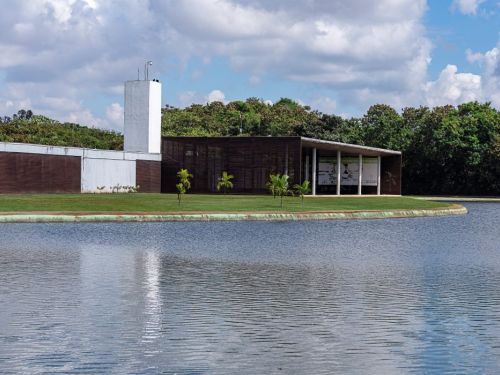 Um edifício moderno à beira do lago, no Parque Ecológico da Pampulha. O prédio é longo e retangular, com paredes escuras e uma torre branca. O primeiro plano é ocupado pela água com pequenas ondas, que reflete o céu. O edifício está cercado por grama verde, com árvores ao fundo e pequenas palmeiras em sua frente.