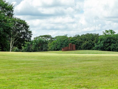 Vasto campo de grama verde claro. No meio do campo, há uma escultura de metal de cor avermelhada, de formato curvo e geométrico. O gramado é delimitado por uma linha de árvores verdes e densas. O céu está cheio de nuvens grandes e brancas.