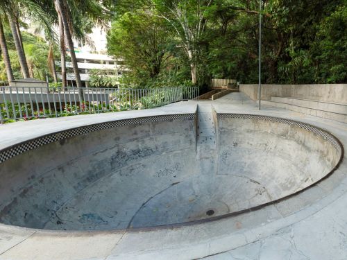 iscina circular vazia de concreto com borda revestida por ladrilhos em preto e branco. O fundo é cinza, com marcas de umidade e um ralo central. A piscina é cercada por corrimão metálico prateado em formato circular. Ao redor há piso cimentado e, além dele, árvores altas e vegetação densa que fecham o espaço com sombra.