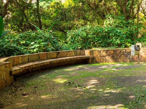 Banco comprido de pedra com encosto baixo, em formato de L, com assento liso e marcas de desgaste. À frente, piso de pedras irregulares, algumas cobertas por musgo verde. No canto direito, lixeira metálica preta está fixada ao chão. Ao fundo, árvores altas e vegetação densa sombreiam o espaço.