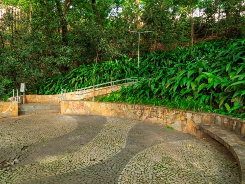 Espaço circular pavimentado com pedras que formam desenhos em curvas e mosaicos. Bancos de pedra, fixos em linha curva, acompanham a forma do espaço. As muretas apresentam grafites em várias cores. Ao fundo, vegetação alta e densa, com árvores fechando o cenário.