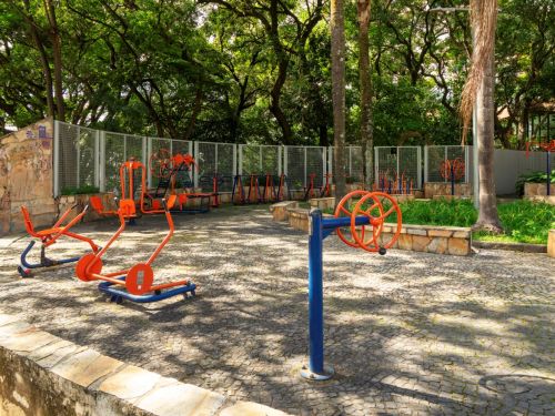 Academia ao ar livre com aparelhos de metal em azul e laranja, fixados sobre piso de pedras irregulares. Equipamentos incluem barras, pedais e bancos. Ao redor, há canteiros de grama e árvores altas que fornecem sombra. O espaço está parcialmente cercado por muretas de pedra.