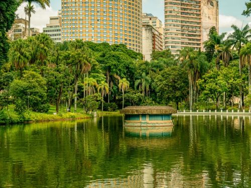 A vista de um lago no Parque Municipal. No meio do lago, há uma pequena construção redonda e azul que se reflete na água verde e ondulada. Atrás das árvores que cercam o lago, surgem dois edifícios altos, um de cor amarela e outro de cor bege. O céu está azul com nuvens brancas.