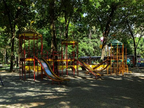 Um parquinho de diversões vazio dentro do parque. O brinquedo é feito de metal amarelo e vermelho, com escorregadores duplos, escadas e plataformas. O parquinho está em uma área de terra com pedrinhas, cercado por muitas árvores grandes e com folhagem densa. A luz do sol passa por entre as copas das árvores, criando um efeito de luz e sombra na cena.