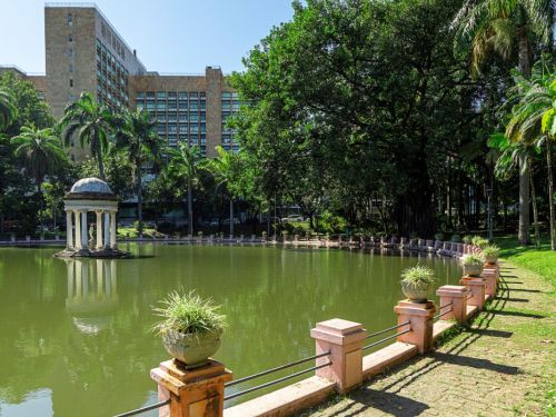 Vista de um lago no Parque Municipal. Em primeiro plano, uma mureta rosa com vasos de plantas divide um caminho do lago. Sobre uma pequena ilha, há um gazebo redondo e branco. As águas verdes do lago refletem a construção e as árvores. No fundo, um grande prédio moderno se mistura com a paisagem, que é repleta de árvores.