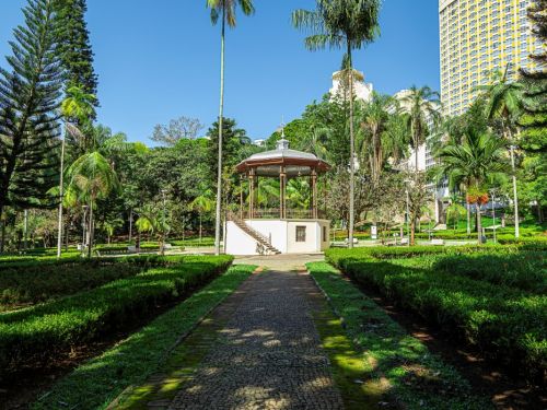 um coreto no parque. Um caminho de pedras escuras se estende em linha reta até a construção, ladeado por pequenos arbustos verdes. O coreto, no fundo, é redondo e branco, com um telhado em cúpula e uma escadaria. A paisagem tem árvores de vários tipos, e um prédio moderno é visível ao fundo, sob um céu azul e sem nuvens.