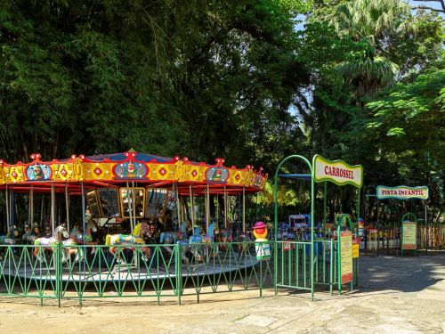 Um carrossel colorido no parque. A atração tem um telhado em vermelho e amarelo, e cavalos e outros animais pintados em cores vibrantes. Uma placa verde indica "CARROSSEL" e "PISTA INFANTIL". A atração está cercada por uma cerca verde e por muitas árvores com folhagem densa, que criam sombras no chão de terra.