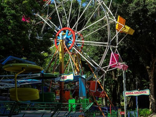 Uma roda gigante no Parque Municipal. A atração tem uma estrutura branca e cadeiras coloridas em tons de vermelho, laranja, amarelo e azul. O letreiro de entrada diz "Roda Gigante". Outras atrações com cores vibrantes estão ao lado. O local é cercado por árvores grandes, com o céu azul visível entre a folhagem.