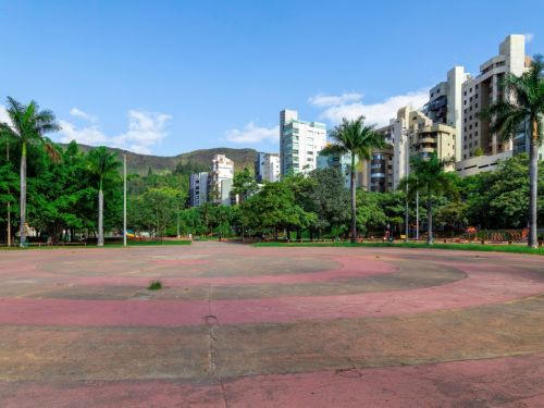 Uma grande praça circular de cimento, com anéis pintados em tons de vermelho desbotado. O local é cercado por palmeiras altas e outras árvores. Ao fundo, vários edifícios residenciais altos se erguem contra um céu azul. Uma serra coberta de vegetação é visível à esquerda.