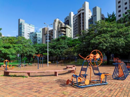 Área de ginástica ao ar livre com piso de tijolos retangulares. Vários aparelhos de metal, em cores azul e laranja, estão dispostos pelo pátio. Há também um banco de cimento de formato curvo. Ao fundo, muitas árvores e diversos edifícios altos sob um céu azul claro.