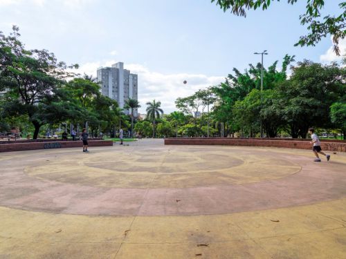 Uma arena circular de cimento liso em tons de amarelo e rosa desbotado. Ao redor, pessoas praticam esportes. O espaço é cercado por um pequeno muro de tijolos e muitas árvores. Ao fundo, um edifício residencial alto se destaca no centro da imagem, sob um céu claro.