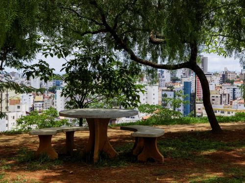 Mesa redonda de concreto com três bancos em um gramado com terra exposta. Árvores ao redor fazem sombra. Ao fundo, muitos prédios brancos e coloridos em uma área urbana.