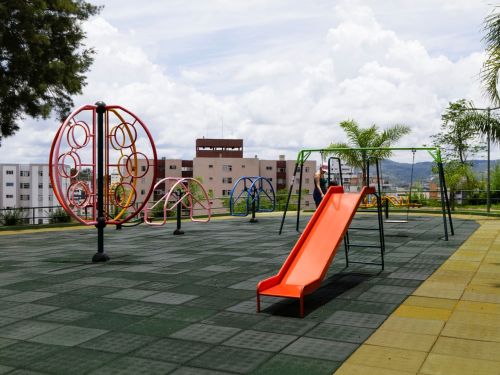Playground com piso emborrachado quadriculado verde e amarelo. Há escorregador laranja, balanços verdes e estruturas metálicas circulares coloridas. Ao fundo, prédios residenciais.