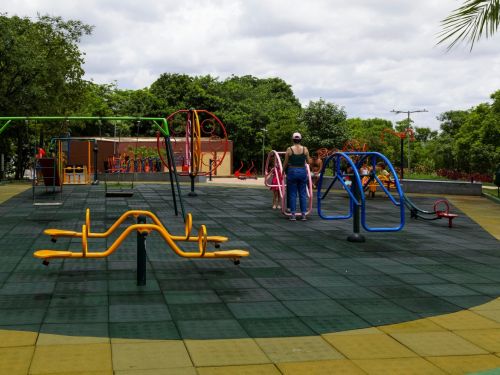 Playground com diversos brinquedos metálicos coloridos sobre piso emborrachado quadriculado. Crianças e adultos utilizam os equipamentos. Árvores verdes cercam a área.