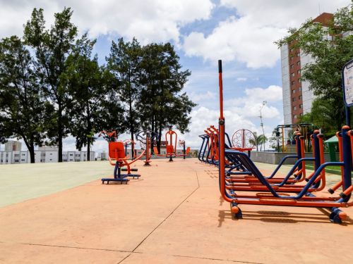 Academia ao ar livre com equipamentos metálicos laranja e azul dispostos lado a lado sobre piso de concreto. Árvores altas ao fundo e edifício residencial ao lado.