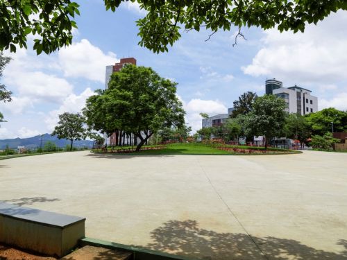 Ampla praça pavimentada em concreto claro, rodeada por árvores e gramado central com canteiros de flores vermelhas. Ao fundo, edifícios residenciais e montanhas.