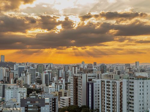 Vista panorâmica de uma cidade com muitos prédios altos. No céu, nuvens escuras deixam passar feixes de luz dourada do pôr do sol, iluminando o horizonte com tons alaranjados.