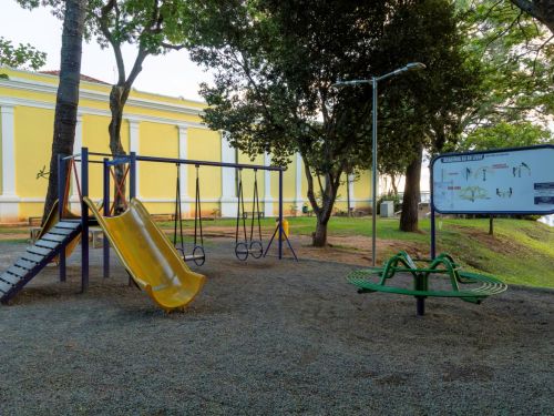 Parquinho infantil com escorregador amarelo, balanços de assento circular, gangorra e brinquedo giratório verde. À direita, placa de academia ao ar livre. Ao fundo, prédio amarelo com colunas brancas e árvores grandes.