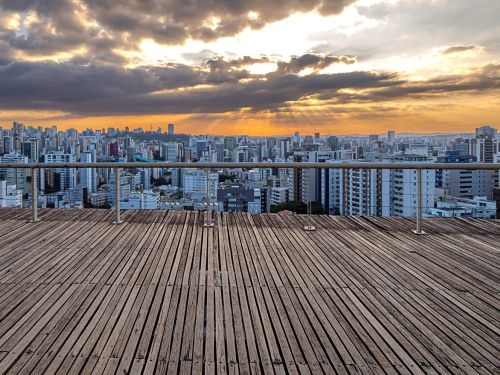 Deck de madeira elevado com corrimão metálico em frente a vista panorâmica da cidade. No horizonte, prédios altos sob céu alaranjado com nuvens escuras e feixes de luz solar atravessando.