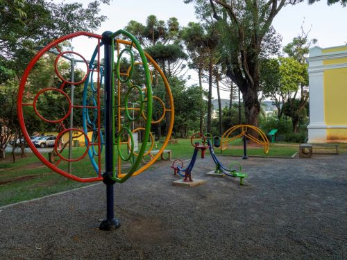 Brinquedos coloridos em parquinho infantil: estrutura circular vertical com argolas, gangorra e equipamentos de exercício. Piso de cascalho. Ao fundo, árvores altas e muro amarelo com base branca.