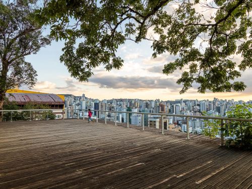 Deck de madeira elevado com corrimão metálico, árvores grandes ao redor e vista panorâmica da cidade ao fundo. Uma criança de blusa rosa caminha próximo ao parapeito.