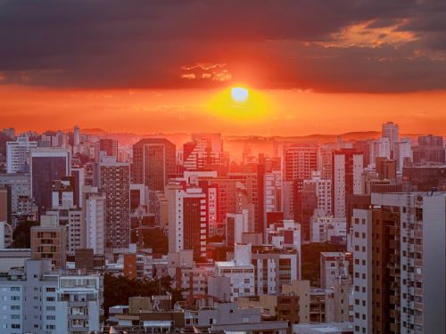 Pôr do sol sobre cidade com muitos prédios altos. O sol alaranjado aparece parcialmente encoberto por nuvens escuras, iluminando a paisagem urbana com tonalidade avermelhada.
