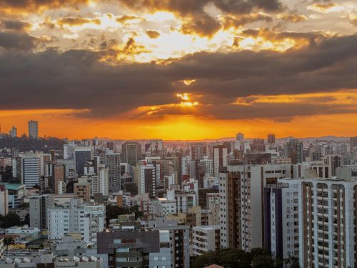Vista panorâmica da cidade com prédios altos. O céu apresenta nuvens escuras e o pôr do sol ilumina o horizonte com tons alaranjados e feixes de luz descendo entre as nuvens.