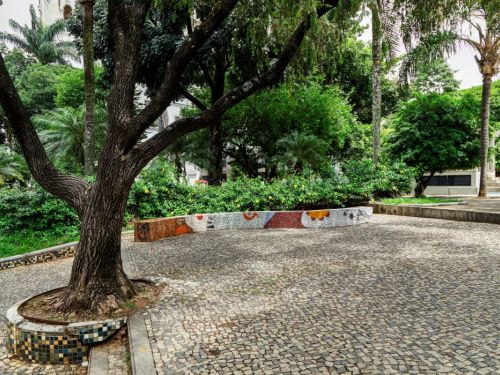 Em primeiro plano, o tronco escuro e robusto de uma árvore emerge de um canteiro baixo, com um piso de pedras portuguesas ao redor. O canteiro, que se estende para a direita, é decorado com um mosaico colorido em tons de branco, laranja e azul. Ao fundo, uma vegetação densa com arbustos e árvores altas.