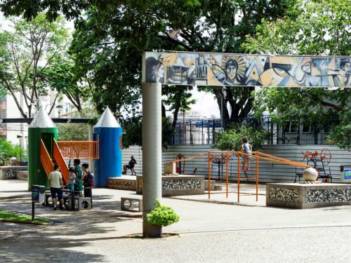 Um parquinho com brinquedos de estrutura metálica e detalhes em plástico colorido. À esquerda, duas torres, uma verde e uma azul, com escorregadores laranja. Ao centro, um painel artístico horizontal e colorido sobre um pilar. Crianças e adultos estão espalhados pelo local, que é cercado por árvores.