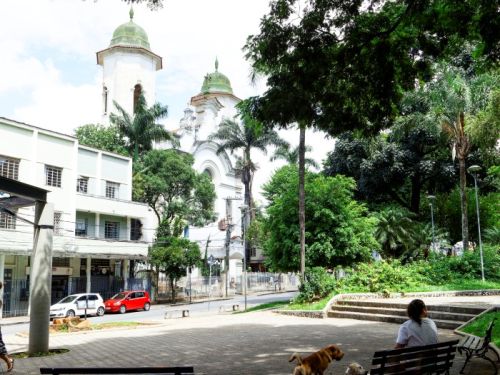 Vista de uma praça a partir de uma área gramada e sombreada por árvores. Uma mulher está sentada em um banco com dois cachorros ao seu lado. À frente, um pátio aberto com mais bancos. Ao fundo, do outro lado da rua, uma igreja branca com duas torres com cúpulas verdes se destaca entre as árvores e edifícios.