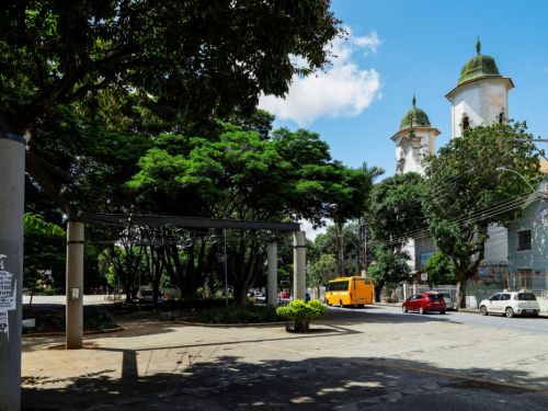 Vista de uma rua a partir da praça. Em primeiro plano, uma área cimentada com pilares de sustentação cinza. À direita, na rua, passam um ônibus escolar amarelo, um carro vermelho e um branco. Ao fundo, entre as árvores, destacam-se as duas torres brancas com cúpulas verdes de uma igreja, sob um céu azul.