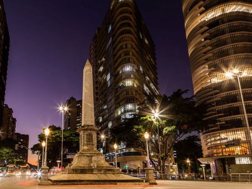 Fotografia noturna com o obelisco em primeiro plano, à esquerda. A cena é iluminada por postes de luz que criam um efeito de estrela. Ao redor, altos edifícios com janelas iluminadas se destacam contra o céu de cor anil. Uma rua com faixas de trânsito circunda o monumento.