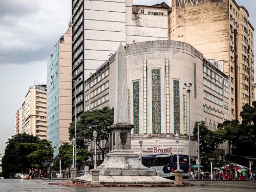 A praça em um dia nublado, com a rua molhada. Em primeiro plano, uma faixa de pedestres com largas listras vermelhas e brancas. Ao centro, o obelisco e, ao fundo, o edifício do Cine Theatro Brasil Vallourec com sua fachada em art déco. Outros prédios altos e de diferentes estilos compõem o entorno.