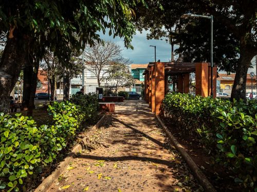 Caminho de terra delimitado por arbustos baixos leva a pergolado de tijolos à vista com vigas de madeira. Postes de iluminação à esquerda. Ao fundo, rua com casas, carros e placas. Piso com folhas espalhadas.
