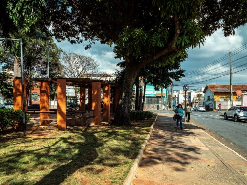 Pergolado de tijolos com vigas de madeira junto à calçada. Área da praça com grama, terra e árvores grandes. Na rua, carros estacionados e pedestres. Postes, fiação e construções baixas ao fundo.