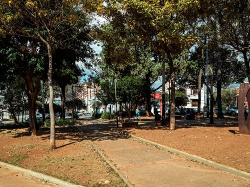 Área aberta com piso de terra e caminhos de concreto cruzados. Árvores espaçadas, bancos e postes ao redor. À direita, escultura metálica ocre em placa vertical oval com abertura central. Construções e carros visíveis ao fundo.