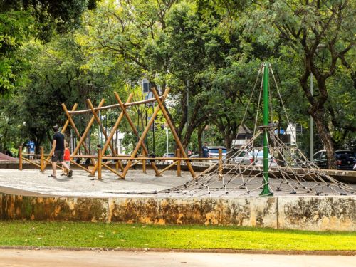Em um parque, uma área de recreação infantil com chão de cascalho claro. À direita, um brinquedo de escalada em formato de pirâmide feito de cordas, com um poste central verde. À esquerda, uma estrutura maior formada por toras de madeira interligadas. Ao fundo, muitas árvores altas e frondosas.