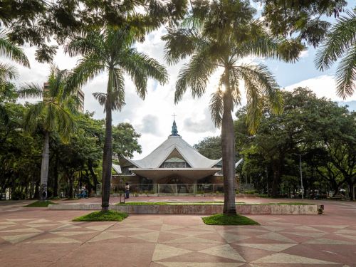Vista frontal de uma igreja de arquitetura moderna em uma praça. O edifício tem um telhado branco de formato parabólico que se eleva a um pico central com uma cruz. A cena é emoldurada por duas palmeiras altas. O piso da praça é de cimento com padrões geométricos em tons de vermelho e creme.