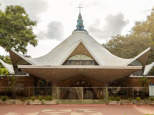 Vista frontal de uma igreja com telhado branco em formato geométrico de grandes abas inclinadas. No topo há uma torre azul com cruz. A fachada possui paredes de tijolos e vitrais triangulares. Em frente, cercas metálicas, vasos com plantas e piso em tons de vermelho e bege. Árvores aparecem nas laterais.