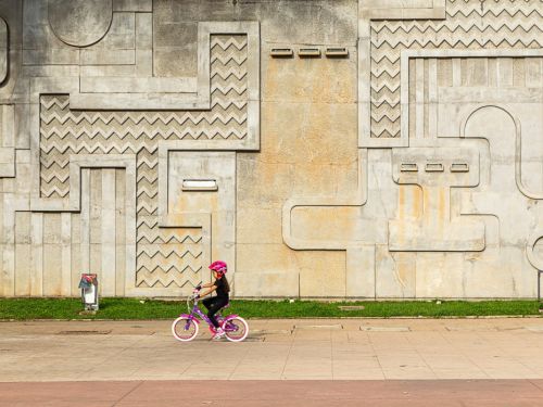 Menina com capacete rosa anda de bicicleta infantil roxa diante de um grande muro de concreto cinza com relevos geométricos. O muro exibe padrões de linhas retas, curvas e zigue-zagues. O chão é pavimentado em bege e vermelho, com faixa estreita de grama verde na base do muro.