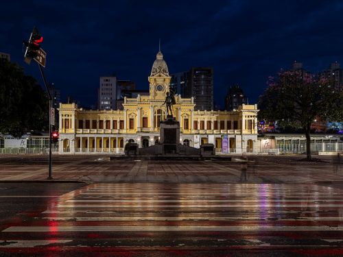 Cena noturna da praça. O edifício da estação está totalmente iluminado em amarelo, destacando-se contra o céu escuro. Em frente, um monumento com uma estátua de bronze no topo. O chão molhado da rua e da faixa de pedestres reflete as luzes dos semáforos e do prédio.